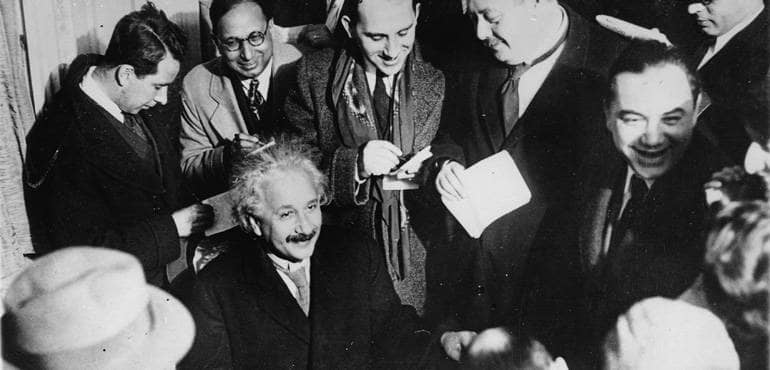 Einstein sitzt und ist von Journalisten umringt.