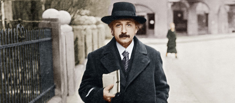 Albert Einstein läuft mit elegantem Hut und Mantel und einem Schriftstück unterm Arm durch eine Straße.